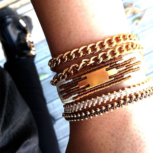 Puede incluir: Una pila de pulseras en la muñeca de una persona. Las pulseras están hechas de cadena dorada, cuerda tejida marrón y blanca, y pequeñas cuentas doradas.