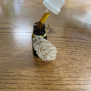 May include: A small brown glass bottle with a white dropper dispensing a golden liquid. A leaf-shaped tag reads "Elixir di Luce" and "For you". The bottle is tied with a gold ribbon. The bottle is on a wooden surface.