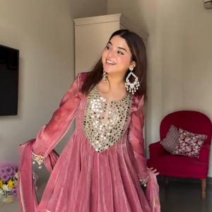 May include: A dusty rose-coloured, long-sleeved dress with intricate mirror and silver embellishments around the neckline. The dress features a flared skirt and matching trousers. The sleeves and trousers are trimmed with silver and white details. The outfit is a traditional style.