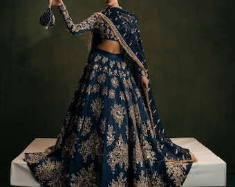 Könnte beinhalten: Ein marineblauer Lehenga mit aufwendiger goldener Stickerei. Das traditionelle indische Kleidungsstück besteht aus einem langen Rock, einem eng anliegenden Oberteil und einem passenden Dupatta. Eine kleine, perlenbesetzte Handtasche wird gehalten. Das Outfit wird mit Kopfschmuck und Ohrringen getragen.