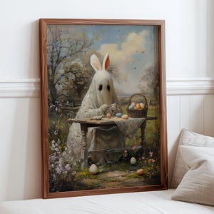 Puede incluir: Una obra de arte enmarcada representa un fantasma con orejas de conejo decorando huevos de Pascua. La escena está ambientada al aire libre con una cesta de huevos y un fondo campestre, enmarcada en madera oscura.