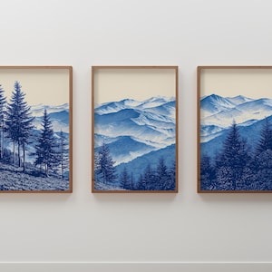 May include: Three framed art prints featuring blue mountain landscapes and evergreen trees. The artwork has a vintage aesthetic with a cream-colored background and brown wooden frames. The prints are displayed on a white wall.