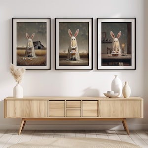 Puede incluir: Tres impresiones artísticas enmarcadas con ilustraciones caprichosas de figuras fantasmales con orejas de conejo. Cada figura sostiene huevos de Pascua en un campo, una cesta o sobre una mesa. Las impresiones tienen una paleta de colores vintage.