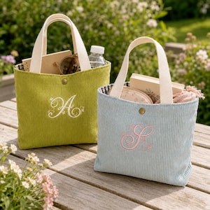 Pode incluir: Duas bolsas tote de veludo cotelê com alças na cor creme e fecho de pressão. A bolsa verde-limão apresenta uma letra A branca bordada, enquanto a bolsa azul-clara exibe uma letra S rosa bordada. Ambas as bolsas contêm objetos em seu interior.