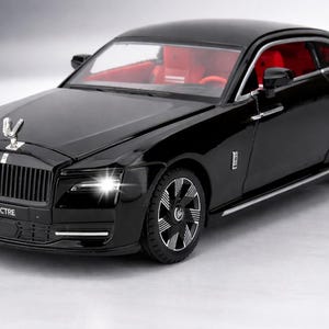 Könnte beinhalten: Ein schwarzes Modellauto mit roter Innenausstattung. Das Auto hat eine silberne Kühlerfigur und das Wort "SPECTRE" auf der Vorderseite. Die Scheinwerfer des Autos sind beleuchtet und die Räder haben ein silbernes und schwarzes Design.