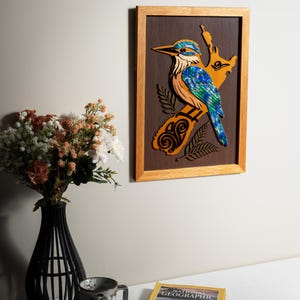 May include: A rectangular wooden wall art piece featuring a layered cutout of a kingfisher bird. The bird is decorated with iridescent blue and green abalone shell inlay, set against a dark brown background within a light oak-coloured wooden frame.