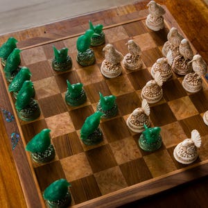 May include: A wooden chess set with a checkered board and bird-shaped pieces. The green pieces are bird-shaped, while the opposing pieces are off-white with intricate details. The board is set on a wooden table.
