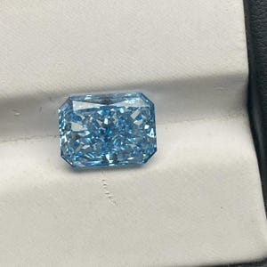 May include: A stunning, rectangular-cut, light blue diamond. The gemstone is presented against a white backdrop within a black box. The diamond's facets catch the light, producing a sparkling effect. The overall impression is one of luxury and sophistication.