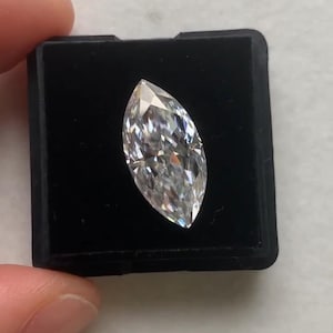 Op de afbeelding: Een marquise-geslepen diamant, gepresenteerd in een zwarte doos. De diamant is helder en facetgeslepen, waardoor het licht wordt gereflecteerd. De vorm is langwerpig met puntige uiteinden. De doos wordt tegen een wit oppervlak gehouden.