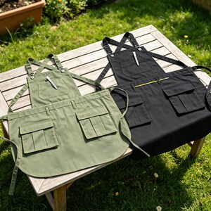 May include: Two aprons, one olive green and one black, rest on a wooden table outdoors. Each apron has multiple pockets and a pen tucked into the chest pocket. The aprons have adjustable straps and waist ties, suitable for cooking or crafting.