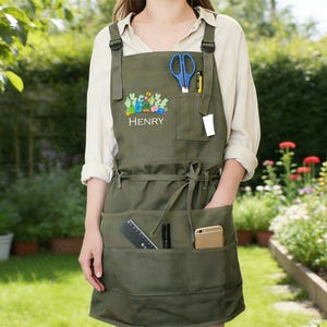 May include: An olive green apron featuring multiple pockets, adjustable shoulder straps, and a waist tie. The chest area displays a colorful potted plant graphic above the name HENRY. Pockets contain blue scissors, a pen, a ruler, and a smartphone.