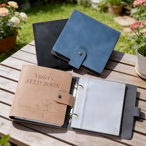 May include: Seed books in tan, blue, and black. The tan book is open, displaying clear plastic sleeves and the text "Violet's SEED BOOK." Each book features a snap closure. The books are on a wooden table.