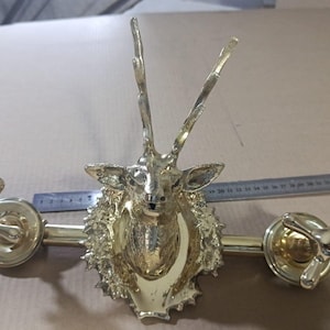 Puede incluir: Decoración de pared de metal dorado con una cabeza de ciervo con grandes astas. La pieza tiene dos pomos decorativos y mide aproximadamente 46 cm de ancho.