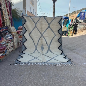May include: A large, off-white rug with a diamond pattern in gray and black. The rug has a fringe along the bottom edge. The rug is displayed outdoors on a paved surface.