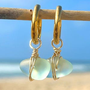 May include: A pair of gold hoop earrings with sea glass pendants. The sea glass is a pale blue-green color, and the hoops are a shiny gold. The earrings are hanging on a wooden stick, with a beach and ocean background.