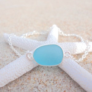 May include: A light blue sea glass pendant necklace with a silver chain. The pendant is oval-shaped and set in a silver bezel. The necklace is displayed on a white starfish, resting on sand.