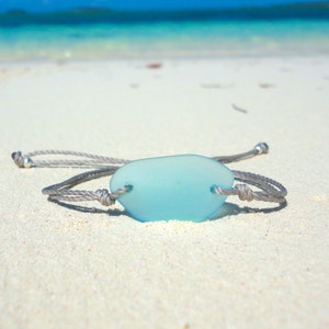 Peut inclure: Un bracelet en corde grise avec un charme en verre de mer bleu clair. Le bracelet est pos&eacute; sur une plage de sable blanc.