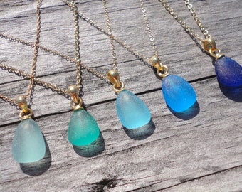 May include: Six gold necklaces with teardrop-shaped sea glass pendants in shades of blue and green. The necklaces are arranged in a row on a wooden surface.