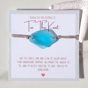 Puede incluir: Un brazalete de vidrio de mar azul claro con un cord&oacute;n gris y una tarjeta que dice "Gracias por ayudarnos a atar el nudo" y "Como el suave vaiv&eacute;n de las olas del oc&eacute;ano, tu apoyo inquebrantable ha significado el mundo para m&iacute;, y estoy profundamente agradecida de tenerte como mi dama de honor."