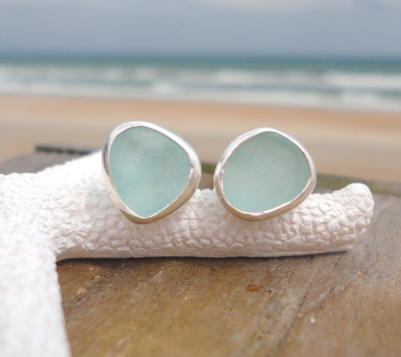 May include: A pair of seafoam green earrings with a silver bezel setting. The earrings have an irregular, organic shape and are displayed on a white, textured surface. The background shows a beach scene with the ocean and sky.
