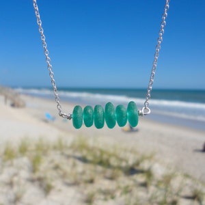May include: A silver chain necklace with a row of five teal sea glass beads. The necklace is photographed against a blurred background of a beach.