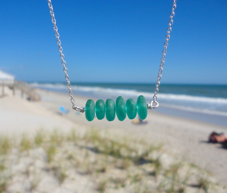 May include: A silver chain necklace with a row of five teal sea glass beads. The necklace is photographed against a blurred background of a beach.