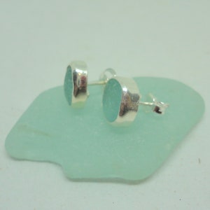 May include: A pair of oval-shaped, seafoam green stud earrings with a silver bezel setting. The earrings are displayed on a light green, irregular-shaped surface. The earrings are small and delicate, perfect for everyday wear.