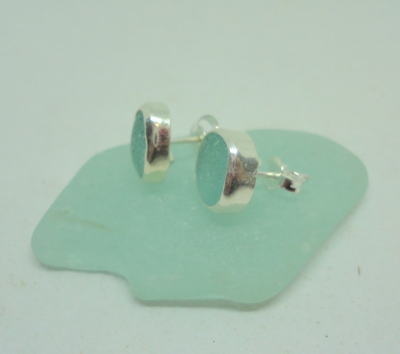 May include: A pair of oval-shaped, seafoam green stud earrings with a silver bezel setting. The earrings are displayed on a light green, irregular-shaped surface. The earrings are small and delicate, perfect for everyday wear.
