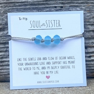 Może przedstawiać: Biała kartka z tekstem "To my Soul Sister" i wiadomością o przypływie i odpływie oceanicznych fal. Kartka ozdobiona jest jasnoniebieskim bransoletką z koralik&oacute;w.