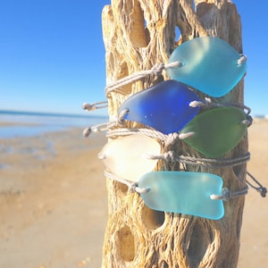 Peut inclure: Cinq bracelets en verre de mer avec un cordon gris, attach&eacute;s ensemble sur un morceau de bois flott&eacute;. Le verre de mer est dans des tons de bleu, vert et blanc.