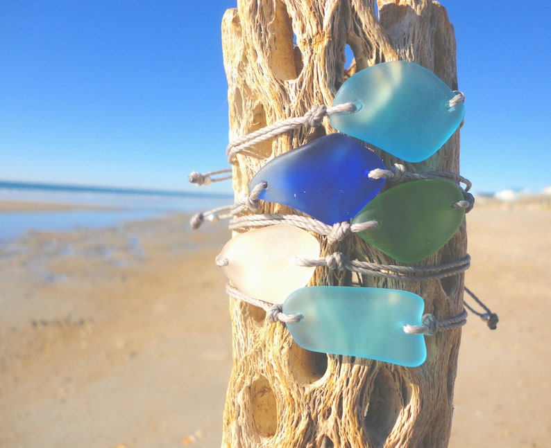 Peut inclure: Cinq bracelets en verre de mer avec un cordon gris, attach&eacute;s ensemble sur un morceau de bois flott&eacute;. Le verre de mer est dans des tons de bleu, vert et blanc.