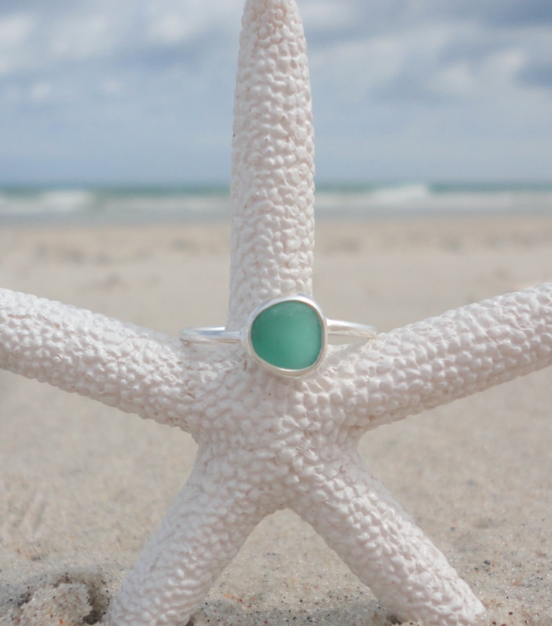 May include: A silver ring with a light green sea glass stone set in a bezel setting. The ring is resting on a white starfish.
