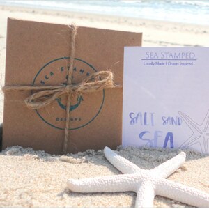 May include: A brown paper package tied with twine and a blue and white logo that says "Sea Stamped Designs". A white card with the text "Sea Stamped Locally Made | Ocean Inspired" and the text "Salt Sand Sea" with a starfish illustration.
