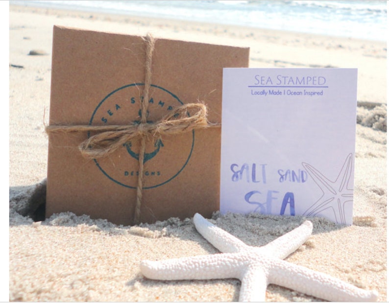May include: A brown paper package tied with twine and a blue and white logo that says "Sea Stamped Designs". A white card with the text "Sea Stamped Locally Made | Ocean Inspired" and the text "Salt Sand Sea" with a starfish illustration.
