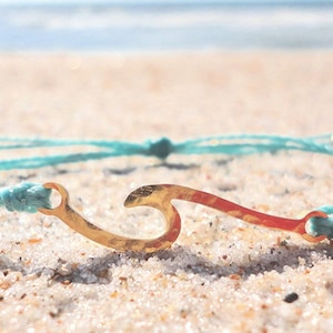May include: A turquoise blue string bracelet with a gold wave charm. The bracelet is lying on a sandy beach.
