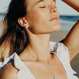 May include: A silver necklace with a teardrop-shaped, light blue pendant. Matching light blue stud earrings are visible. The person is wearing a white top with ruffled sleeves. A silver ring is on the person's finger.