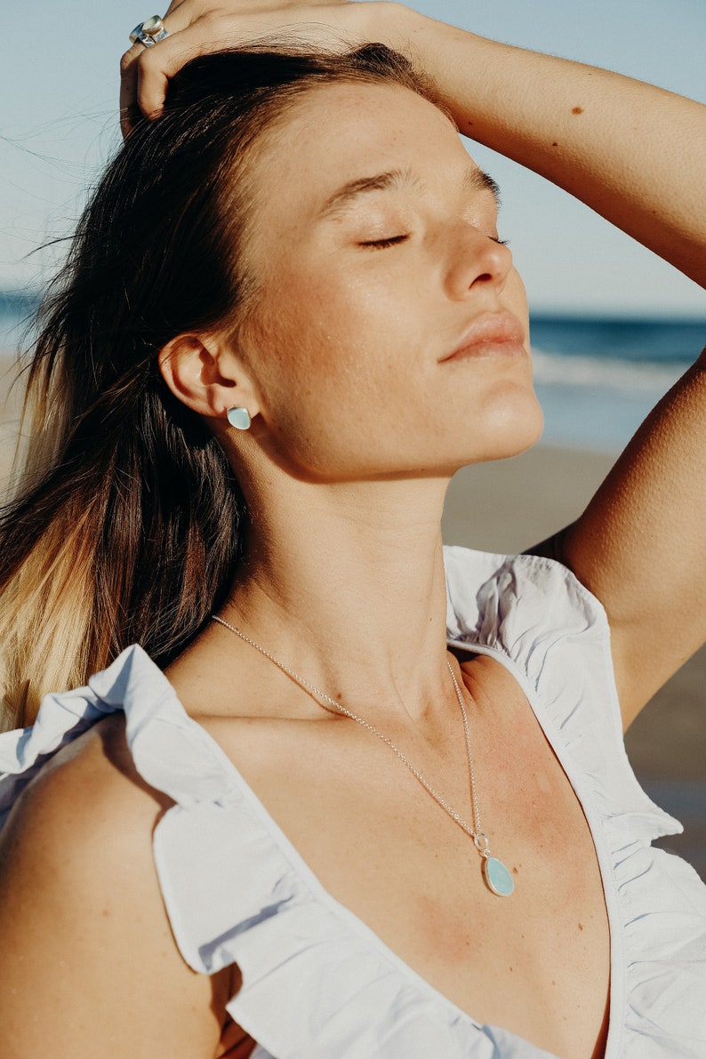 May include: A silver necklace with a teardrop-shaped, light blue pendant. Matching light blue stud earrings are visible. The person is wearing a white top with ruffled sleeves. A silver ring is on the person's finger.