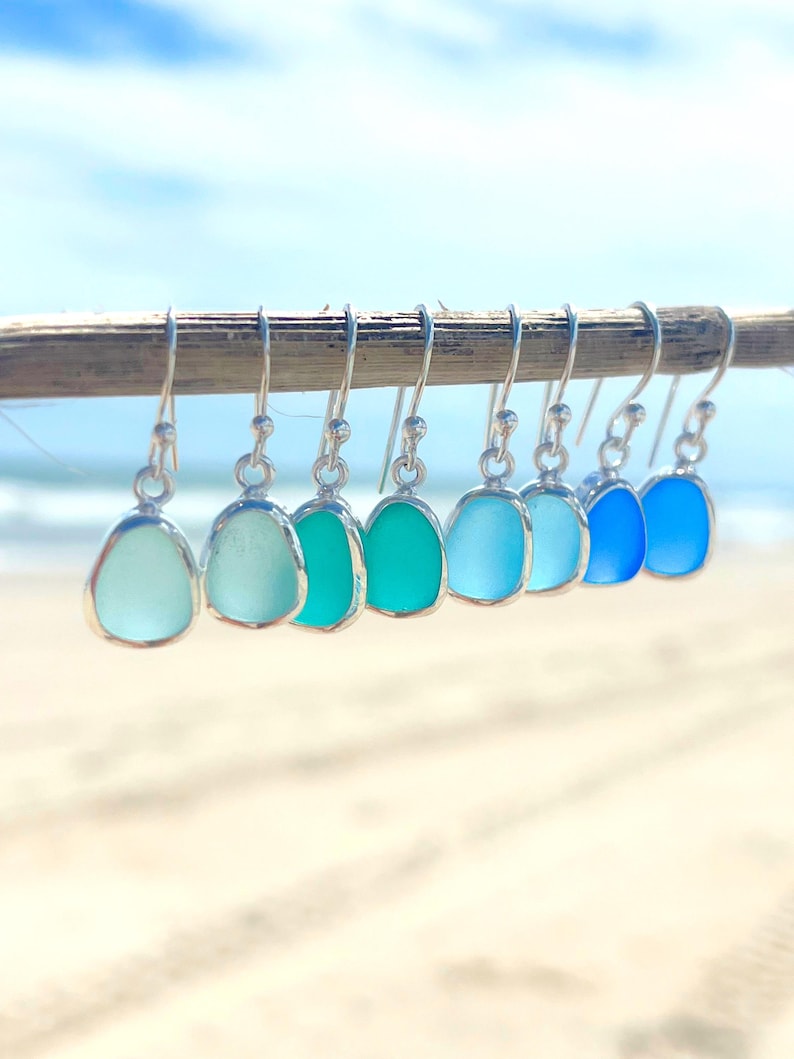 May include: A collection of seven sea glass earrings in varying shades of blue and green, displayed on a wooden stick. Each earring features a silver-colored bezel setting and a hook closure. The background shows a beach and the ocean.
