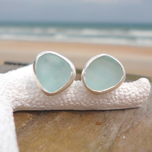 May include: A pair of sea glass stud earrings. Each earring features a light blue, irregularly shaped piece of sea glass set in a silver bezel. The earrings are displayed on a white, textured surface with a blurred beach background.