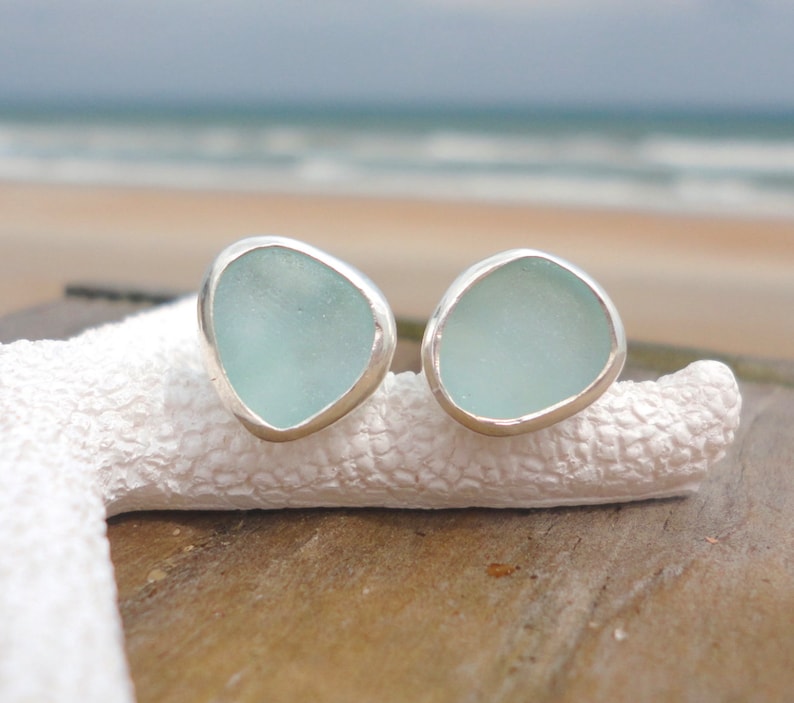 May include: A pair of sea glass stud earrings. Each earring features a light blue, irregularly shaped piece of sea glass set in a silver bezel. The earrings are displayed on a white, textured surface with a blurred beach background.