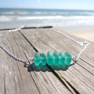 May include: A silver chain necklace with a row of six teal sea glass beads. The necklace is on a weathered wooden surface.