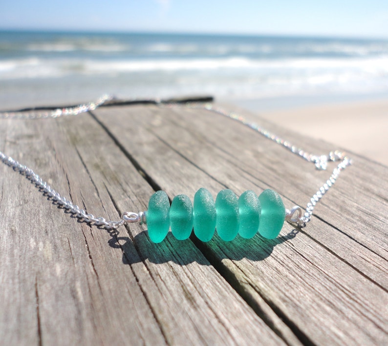 May include: A silver chain necklace with a row of six teal sea glass beads. The necklace is on a weathered wooden surface.