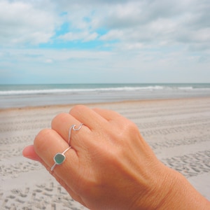 May include: A hand wearing two silver rings on a finger. The ring on the index finger is a simple wave design. The ring on the middle finger is a square-shaped ring with a light green stone.