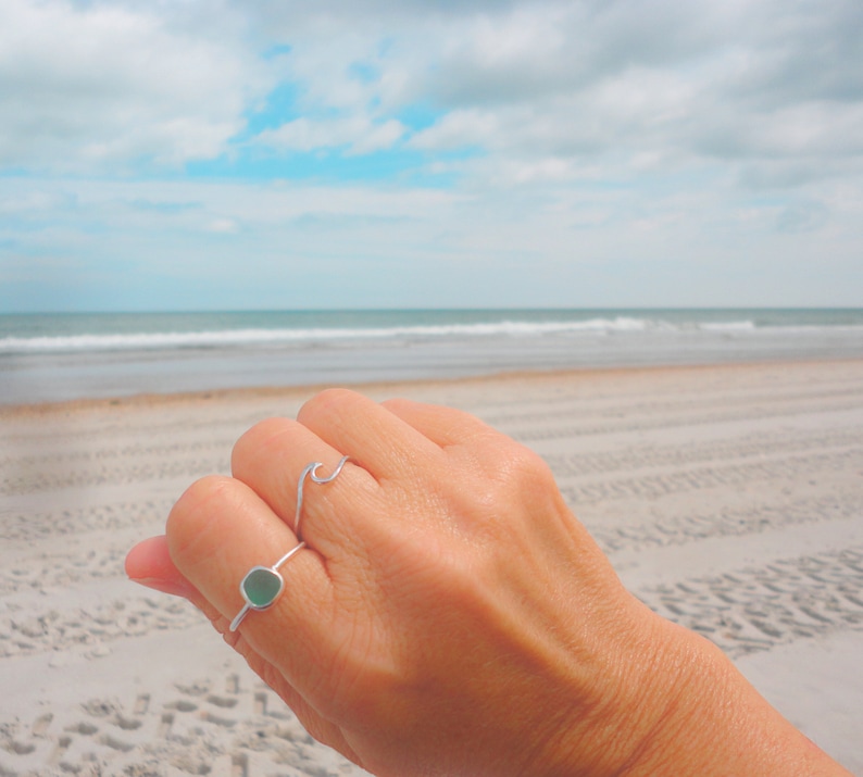 May include: A hand wearing two silver rings on a finger. The ring on the index finger is a simple wave design. The ring on the middle finger is a square-shaped ring with a light green stone.