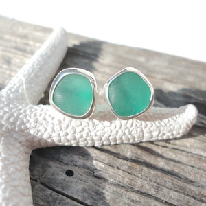 May include: A pair of sea glass stud earrings. Each earring features a piece of teal sea glass set in a silver bezel. The earrings are displayed on a white starfish and a weathered wooden surface.
