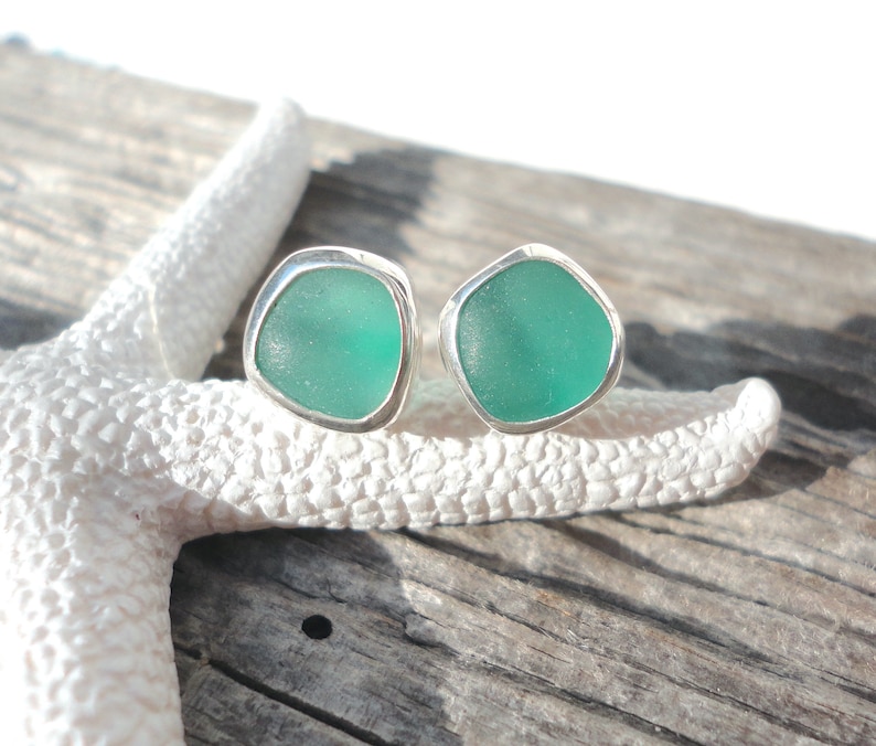 May include: A pair of sea glass stud earrings. Each earring features a piece of teal sea glass set in a silver bezel. The earrings are displayed on a white starfish and a weathered wooden surface.