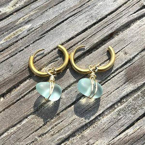 May include: A pair of gold-colored hoop earrings, each with a light blue, heart-shaped sea glass pendant. The sea glass is wrapped in gold-colored wire. The earrings are displayed on a weathered, wooden surface.