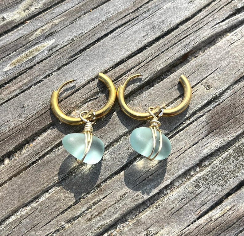 May include: A pair of gold-colored hoop earrings, each with a light blue, heart-shaped sea glass pendant. The sea glass is wrapped in gold-colored wire. The earrings are displayed on a weathered, wooden surface.