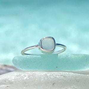 May include: A silver ring featuring a square-shaped sea glass stone. The ring is resting on a piece of sea glass.