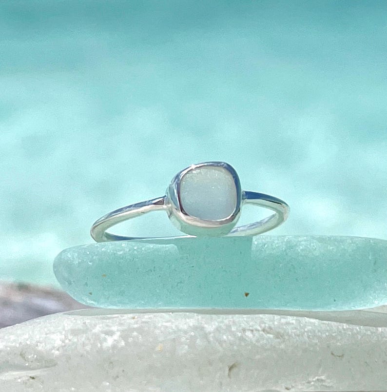 May include: A silver ring featuring a square-shaped sea glass stone. The ring is resting on a piece of sea glass.